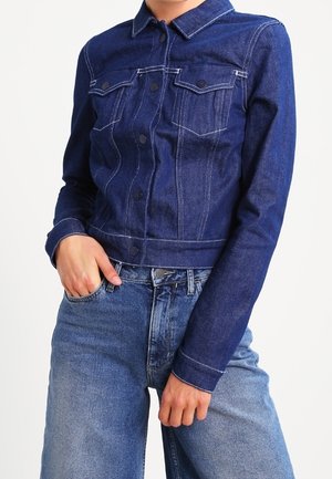 Personne portant une veste en jean bleu foncé avec boutons et un jean taille haute bleu clair, une main dans la poche et l'autre posée sur la cuisse.