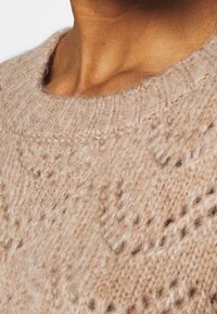 Suéter de punto color beige con un cuello redondo, que presenta un patrón texturizado y un hilo suave y peludo. Primer plano que muestra el detalle de la tela.