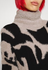 Il maglione presenta una consistenza morbida e pelosa con un collo alto. Il design include modelli astratti in contrasto tra nero e beige chiaro.