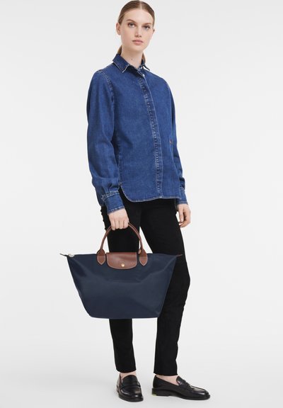 Navy nylon tote bag featuring brown leather handles, a gold snap button, and a minimalist design; accompanied by a denim jacket and black trousers.