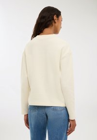 Kremowy sweter o luźnym kroju, z żeberkowym okrągłym dekoltem i opuszczonymi szwami ramion, zestawiony z niebieskimi dżinsami.