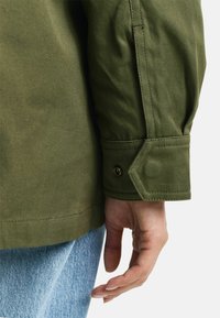 Olivgrüne Jacke mit einem geknöpften Manschette, die einen robusten Stofftextur und eine taillierte Passform aufweist, kombiniert mit hellblauen Jeans.