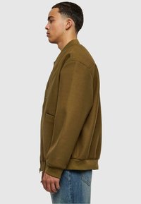 Olivenfarget bomberjakke i myk stoff, med glidelåslukking, sidelommer og ribbestrikkede mansjetter, paired med lyseblå jeans.