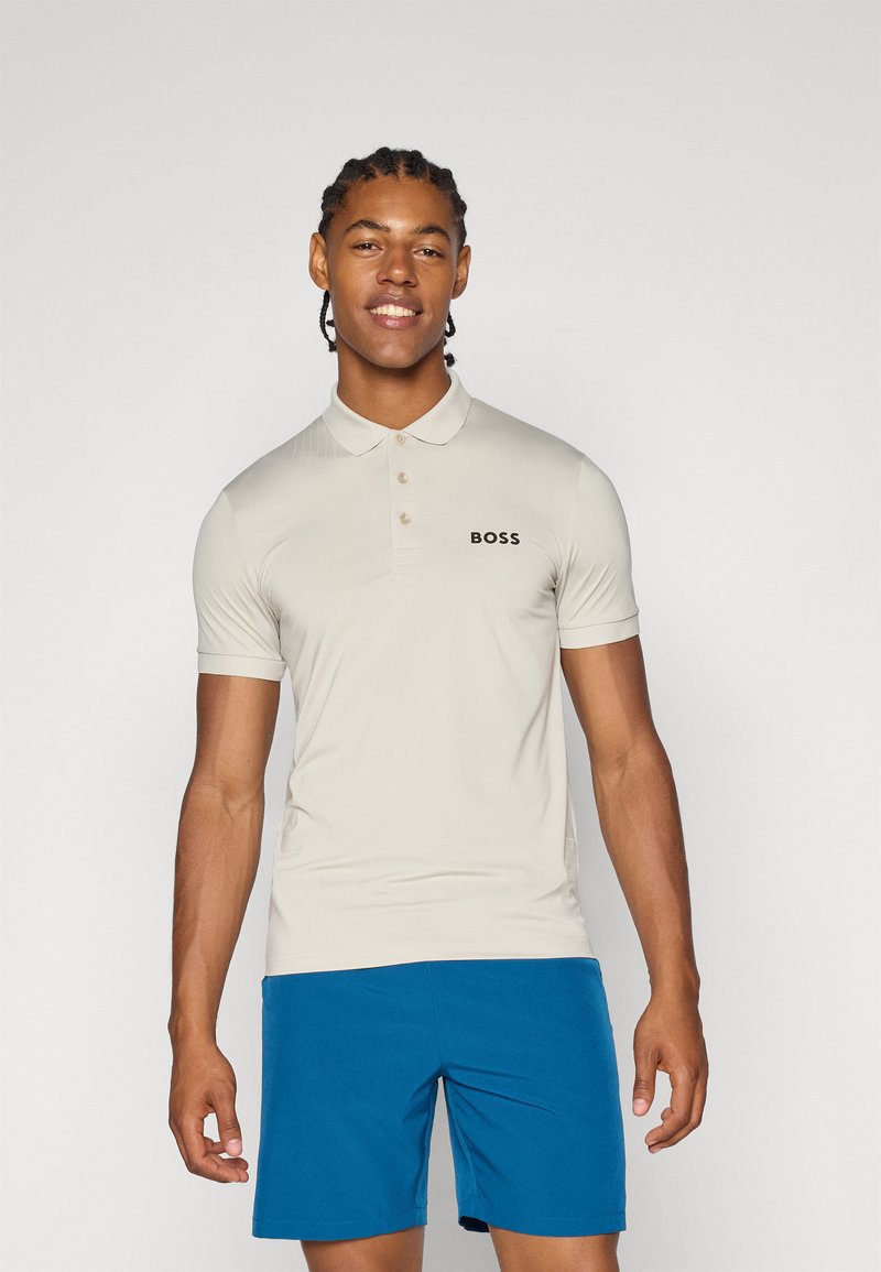Polo beige a maniche corte con tre bottoni, caratterizzato da un logo nero "BOSS" sul petto sinistro. Indossato con pantaloni corti blu.