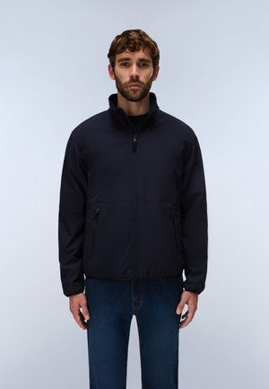 Homme portant une veste noire zippée avec des poches latérales zippées et un jean bleu foncé, debout devant un fond clair uni.