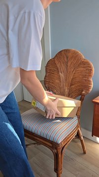 Pacote de cartão segurado com ambas as mãos, colocado numa almofada de listras de uma cadeira de madeira com um encosto em forma de leque.