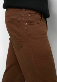 Close-up van een persoon die een bruine broek draagt met zichtbare stiksels en klinknagels op de achterzak, gecombineerd met een zwart topje.