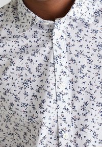 Chemise blanche boutonnée arborant un motif floral avec des accents bleus et rouges, un col pointu et une texture lisse.