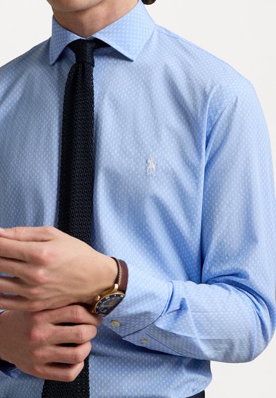 Chemise button-up bleu clair avec un motif blanc subtil, manches longues et un petit logo sur la poitrine ; associée à une cravate en tricot sombre.