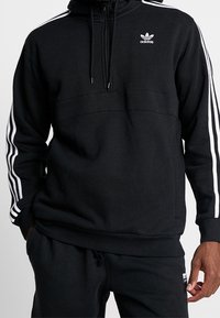 Svart huppartröja med blixtlås framtill, med vita tre-randiga detaljer längs ärmarna och en liten Adidas-logotyp på bröstet.