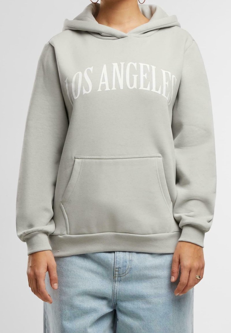 Personne portant un sweat à capuche gris clair avec le texte "LOS ANGELES" sur le devant et un jean bleu clair, main avec bague visible sur le côté.