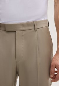 Beige trousers met een gladde textuur, uitgerust met een platte voorkant, dunne riemlussen en subtiele stiksels langs de naden.