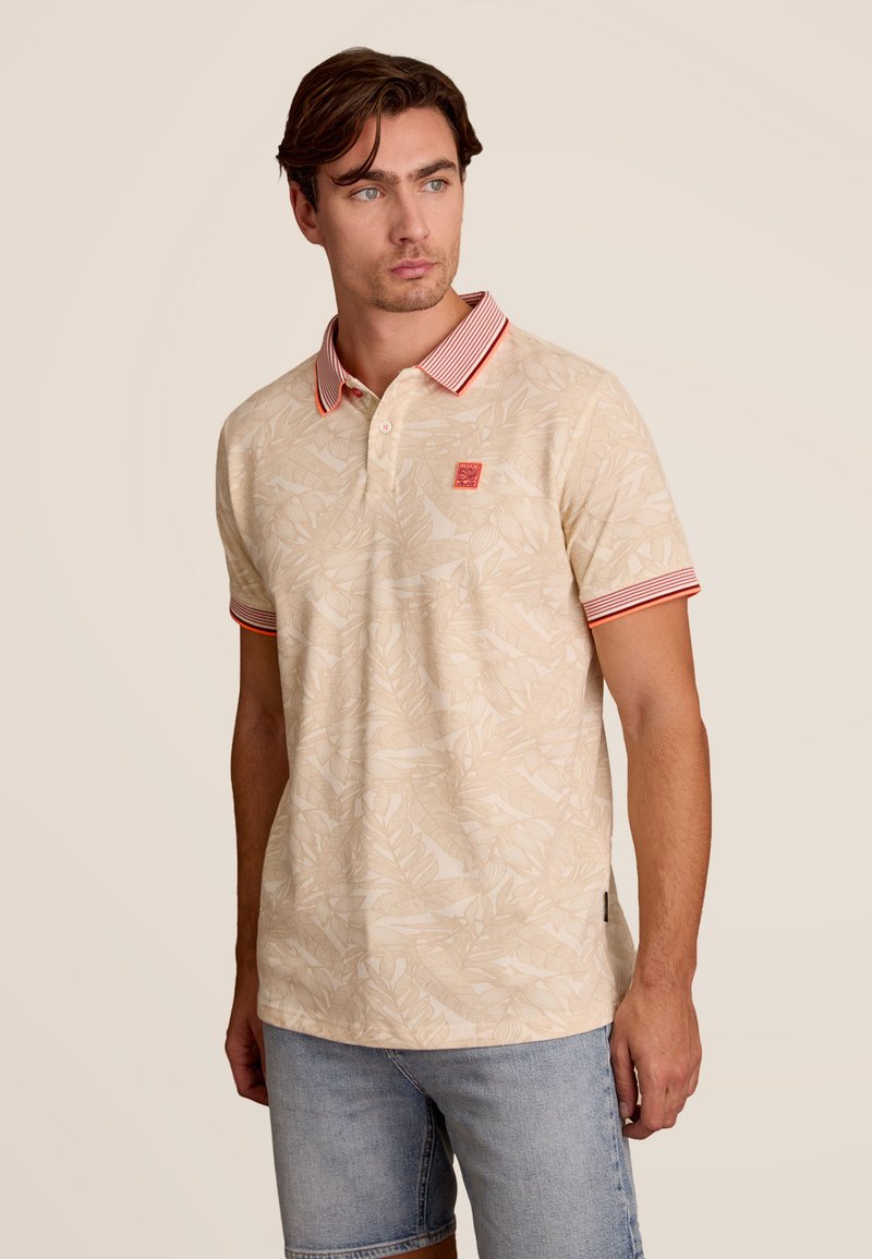 Beige polo shirt met bladdessin, gestreepte rode kraag, twee knoopsluiting en een klein logopatch op de borst. Draagt met denim shorts.