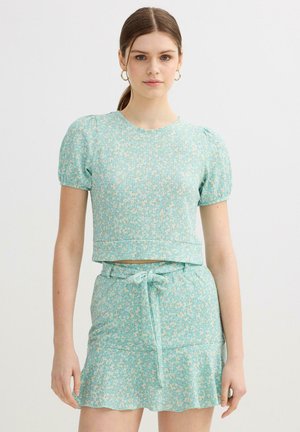 Mujer joven que lleva una blusa de manga corta floral azul clara y una falda de talle alto a juego con un cinturón atado, de pie frente a un fondo sencillo.