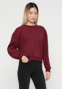 Ung kvinna med rakt ombre-hår, iförd en vinröd broderad sweatshirt med ärmar och svarta tighta jeans, ståendes med ena handen i fickan.