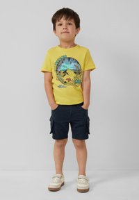 Geel katoenen t-shirt met een kleurraf grafiek van bergen en palmbladeren, gecombineerd met marineblauwe cargoshorts en lichtgekleurde sneakers.