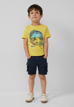 Geel katoenen t-shirt met een kleurraf grafiek van bergen en palmbladeren, gecombineerd met marineblauwe cargoshorts en lichtgekleurde sneakers.