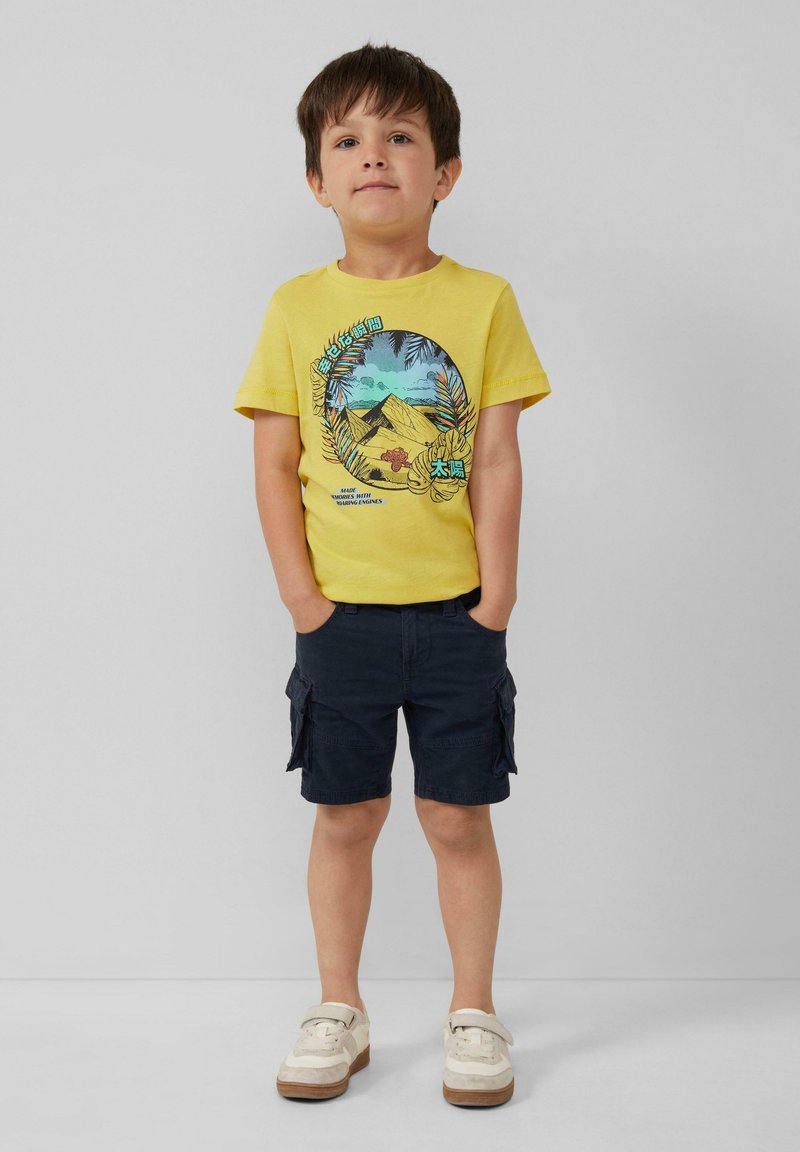 Geel katoenen t-shirt met een kleurraf grafiek van bergen en palmbladeren, gecombineerd met marineblauwe cargoshorts en lichtgekleurde sneakers.