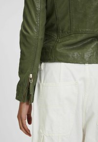 Olivengrüne Lederjacke mit sichtbaren Nähten, einer taillierten Passform und einem Reißverschluss am Ärmel, kombiniert mit weißen Cargo-Hosen.