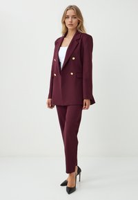 Bordeaux blazer op maat met dubbele gouden knopen, gecombineerd met bijpassende tapered broek. Gestyled met zwarte stilettohakken.
