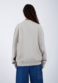 Pullover oversized grigio chiaro con orlo e polsini a costine, caratterizzato da una texture morbida. Abbinato a jeans in denim blu a gamba larga.