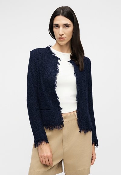 Γυναίκα που φοράει ένα ναυτικό μπλε υφασμάτινο σακάκι με υφή και κρόσσια στο τελείωμα πάνω από λευκό τοπ και μπεζ σορτς ψηλόμεσα, που στέκεται μπροστά σε απλό φόντο.