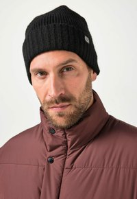 Schwarze Strickmütze mit gerippter Textur, getragen von einer Person in einer kastanienbraunen Daunenjacke mit Knöpfen am Kragen.