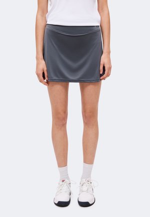 Mujer con falda deportiva gris oscuro, calcetines blancos hasta el tobillo, zapatillas blancas y camiseta blanca, de pie frente a un fondo liso.