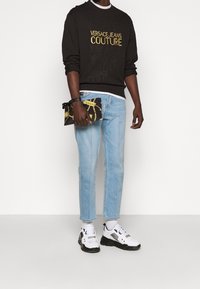 Černá mikina s zlatým logem "Versace Jeans Couture", světle modré džíny a bílé tenisky s černými akcenty. Drží vzorovanou kabelku.