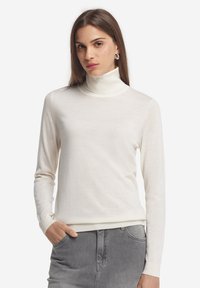 Weißer Rollkragenpullover aus weichem Material, mit langen Ärmeln und gerippten Bündchen. Getragen mit grauen Jeans, die ein schlichtes Design zeigen.