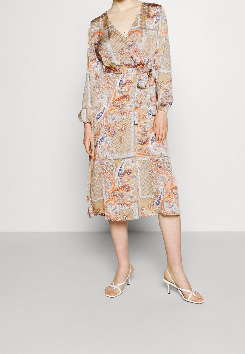 Femme portant une robe portefeuille midi beige avec un motif paisley multicolore et des sandales à talons blanches, debout devant un fond uni.
