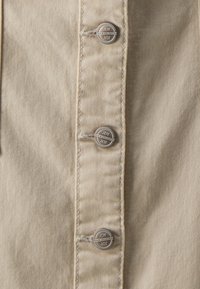 Vaaleanruskea farkkupaita, jossa on viisi metallista napista etupaneelissa, näkyvillä ompeleilla ja sileällä kankaan pinnalla.