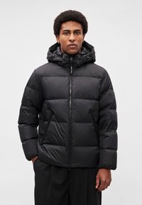 Veste zippée noire matelassée avec capuche, dotée d'un design matelassé, fermeture éclair et poches latérales. Le matériau semble lisse et léger.