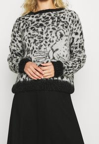 Frau in einem schwarz-weißen, flauschigen Pullover mit einem Leopardenmuster, kombiniert mit einem schwarzen Rock, die die Hände vor der Brust gefaltet hat.