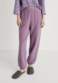 Violet-coloured verryhousut, jotka on valmistettu pehmeästä kankaasta ja joissa on joustava vyötärö sekä kuminauhalla viimeistellyt nilkat, yhdistettynä harmaisiin suoraksi liu'utettaviin tossuihin.