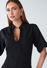 Črna obleka z visokim ovratnikom in kratkimi rokavi. Ima globok V-izrez in gladko teksturo. Značilna je po čistem, minimalističnem dizajnu.