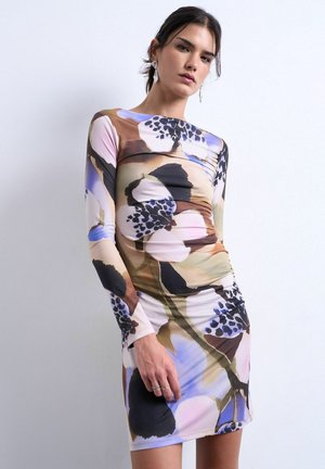 Vrouw die een nauwsluitende jurk met lange mouwen draagt, bedrukt met grote abstracte bloemenpatronen in beige, zwart, blauw en bruintinten, tegen een eenvoudige lichte achtergrond.