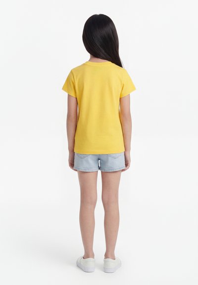 Enfant aux longs cheveux foncés portant un t-shirt jaune à manches courtes, un short en denim clair et des baskets blanches, debout dos à un fond blanc.