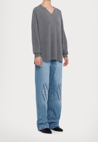 Pull gris en maille avec un large col en V et une texture côtelée, associé à un jean en denim bleu clair avec des accents de logo bleu foncé. Chaussures noires pointues.
