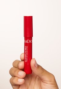 Bastoncino per labbra rosso a matita con finitura lucida. Presenta testo bianco sul tubo, tenuto in una mano con smalto per unghie neutro.