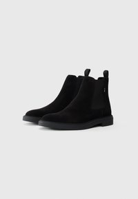 O pereche de botine Chelsea negre din suede, cu panouri elastice laterale și bucle de prindere, expuse pe un fundal gri deschis simplu.