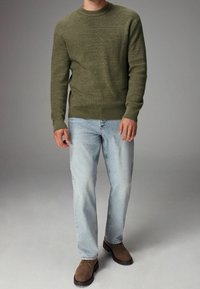 Man in een olijfgroene gebreide trui, lichtblauwe jeans en bruine suède laarzen, staand tegen een effen grijze achtergrond.