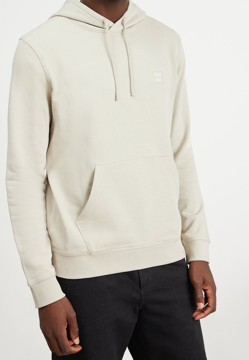 Beige hoodie tillverkad av mjuk bomullsblandning, med en känguruficka, justerbar dragsko huva och diskret logopatch på bröstet.