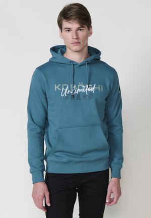 Felpa teal con cordini e tasca frontale; presenta il testo stampato "KOLÖN" e "Unlimited Brand." Polsini e orlo a coste, texture liscia.