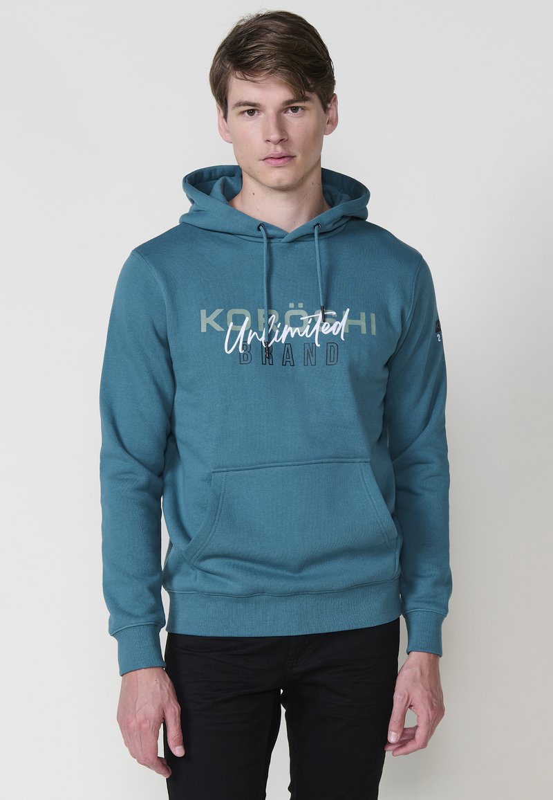Felpa teal con cordini e tasca frontale; presenta il testo stampato "KOLÖN" e "Unlimited Brand." Polsini e orlo a coste, texture liscia.