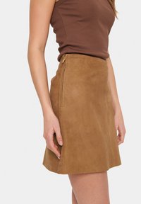 Jupe A-line en suède marron avec une fermeture éclair latérale et une texture lisse. La jupe présente une silhouette épurée et est montrée sur le côté d'un mannequin.
