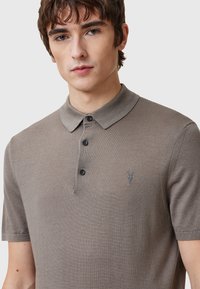 Brun kortärmad polo skjorta med krage, tre knappar och en broderad detalj på vänster bröst. Mjuk, texturerad tyg.