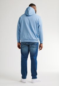 Světle modrá mikina s klokaní kapsou a šňůrkovou kapucí, spárovaná s modrými džíny a bílými teniskami, viděná ze zadní strany.