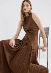 Bruine mouwloze jurk met een aangesloten lijfje, geplooide rok en een ceintuur in de taille. Gladde stof met een subtiele textuur.