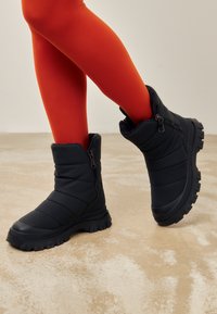 Zwarte puffer enkellaarsjes met een gestructureerde bovenkant, zijrits en dikke rubberen zool met diepe profiel voor grip. Draagt met felrode leggings.